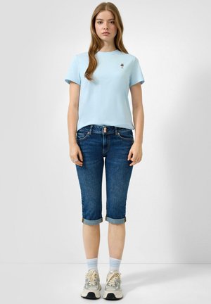 Junge Frau trägt ein hellblaues T-Shirt mit kleinem besticktem Design, hochgekrempelte dunkelblaue Jeansshorts, weiße Socken und beige Sneakers.