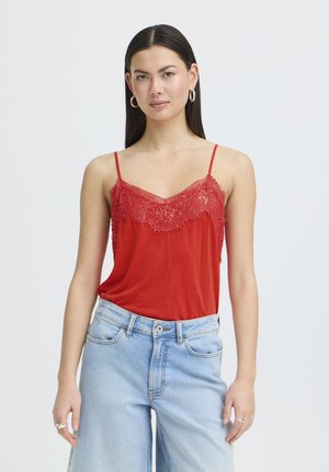 Femme aux longs cheveux foncés portant un caraco rouge avec bordure en dentelle et un jean taille haute bleu clair, debout devant un fond uni.