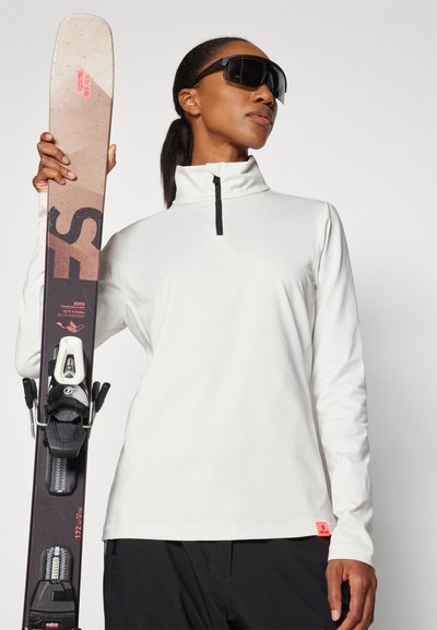 Λευκή μπλούζα με μακριά μανίκια, ψηλό γιακά και μαύρο φερμουάρ, συνδυασμένη με σκούρες σαλοπέτες σκι; το άτομο κρατά ένα σκι με σχέδιο.