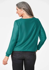 Tealfarbene Bluse mit langen Ärmeln, lässiger Passform und rundem Ausschnitt. Glattes Material mit einem leichten Glanz; kombiniert mit dunkelblauen Jeanshosen mit dekorativen Nähten.