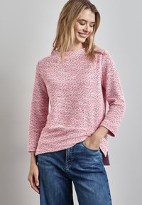 Rosa strukturiertes Sweatshirt mit hochgeschlossenem Kragen und Dreiviertelärmeln, kombiniert mit blauen Jeans. Das Sweatshirt hat seitliche Schlitze für mehr Bewegungsfreiheit.