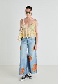 Top sem mangas com padrão verde e amarelo, jeans de perna larga azuis com estampas de hibisco laranja, ténis pretos e uma mala de macramé.