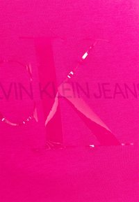Fuchsiová látka s lesklým designem "OK" a textem "CALVIN KLEIN JEANS" v matném provedení, vytvářející texturovaný kontrast.