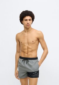 Szürke és fekete úszónadrág, húzózsinóros derékrésszel és oldalzsebbel. Az anyag sima, könnyű textúrájú.