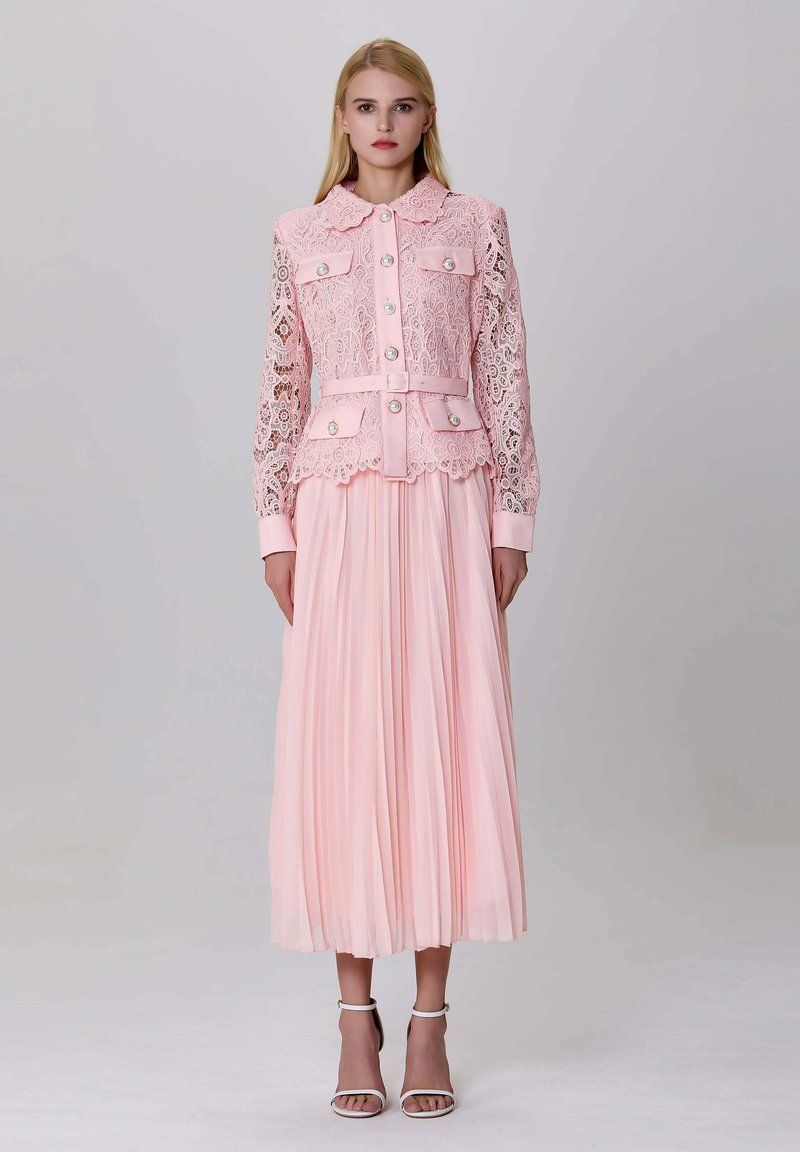 Femme portant une veste en dentelle rose boutonnée avec des accents en perles, une jupe midi plissée assortie et des escarpins blancs à bride de cheville, se tenant sur un fond gris.