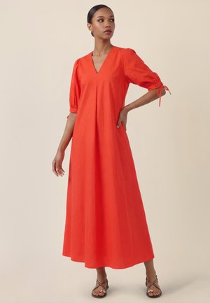 Woman wearing a bright orange V-neck midi dress with puffed elbow-length sleeves and tie details, paired with brown strappy sandals.