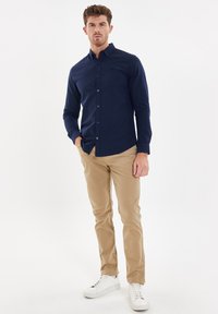 Marineblauwe lange mouwen shirt met button-down kraag en subtiele borstlogo, gecombineerd met beige broek en witte sneakers, staand tegen een witte achtergrond.
