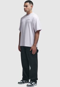 Lys lilla oversized bomulls t-skjorte med korte ermer, med en liten sydd logo på brystet, kombinert med svarte baggy bukser og joggesko.