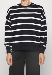 Sudadera negra con rayas horizontales blancas, cuello redondo y mangas largas. Tejido suave con puños y dobladillo acanalados.