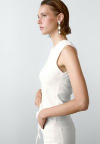 Witte mouwloze top met een getailleerd ontwerp en ronde hals. Gladde textuur. Het model draagt het in combinatie met een broek met een trekkoord in de taille.