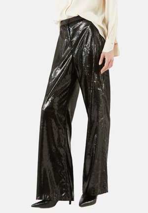 Pantaloni larghi neri lucidi realizzati in tessuto testurizzato, con chiusura frontale e vita alta, abbinati a una maglia di colore chiaro.