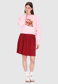 Sudadera corta de color rosa claro con estampado gráfico, combinada con una falda plisada roja hasta la rodilla. La modelo lleva calcetas blancas y zapatillas deportivas.