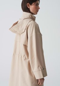 Impermeabile beige con cappuccio, caratterizzato da una vestibilità comoda, grandi tasche laterali e polsini a pressione. Realizzato in materiale morbido e leggero.