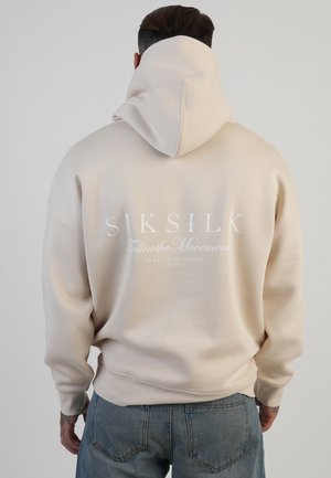 Beige hoodie met een grote capuchon, voorzien van witte bedrukte tekst op de achterkant. Zachte stof textuur en geribbelde manchetten aan de mouwen.