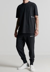 Sort bomulds T-shirt med korte ærmer og et lille hvidt logo på fronten, parret med sorte joggerbukser med et hvidt "genanvendt" logo.