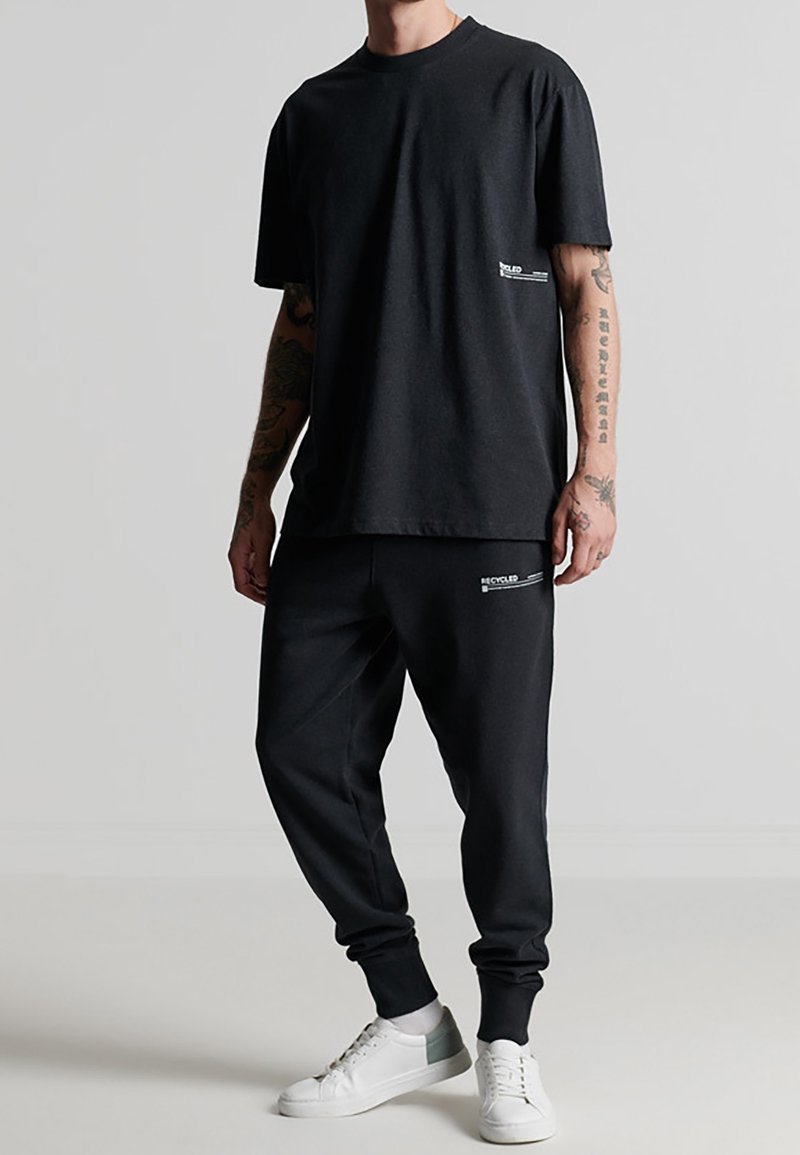 Sort bomulds T-shirt med korte ærmer og et lille hvidt logo på fronten, parret med sorte joggerbukser med et hvidt "genanvendt" logo.