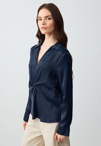 Navy Satinbluse mit V-Ausschnitt, langen Ärmeln und einem geknoteten Design an der Taille. Glatte Textur mit subtiler Sheen.