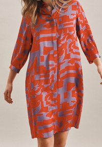 Mujer vestido de manga larga a la altura de la rodilla con un patrón geométrico abstracto en rojo y púrpura sobre un fondo neutro.