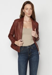 Giacca in pelle marrone con finitura testurizzata, vestibilità slim, chiusura con zip e dettagli in bottoni. Abbinata a un top beige e jeans blu scuro.