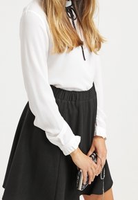 Kvinde iført en hvid langærmet bluse med sort bånd og sort nederdel, der holder en lille clutch med sort-hvidt mønster.