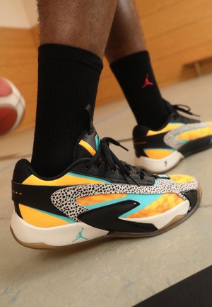 Persona con calcetines negros de Jordan y zapatillas de baloncesto Air Jordan coloridas, de pie en una cancha cubierta con una pelota de baloncesto visible.