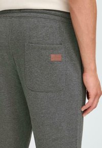 Grå bomulls joggebukser har en enkelt baklomme med et brunt lærmerke. Stoffet er mykt med en subtil tekstur.