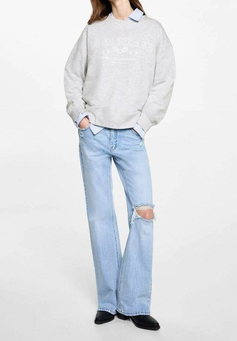 Femme portant un sweat-shirt gris "Long Island" par-dessus une chemise à col bleue, un jean évasé bleu clair déchiré, et des chaussures noires, debout les mains dans les poches.