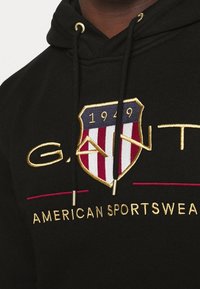 Sudadera negra con capucha que presenta un logo de escudo con franjas rojas y blancas, detalles en dorado, y "GANT" y "AMERICAN SPORTSWEAR" bordados en dorado.