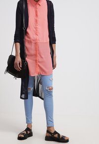 En rosa skjorta med knappar under en svart lång kofta, som kombineras med ljusblå, ripped skinny jeans och svarta sandaler med remmar. En svart handväska ingår.