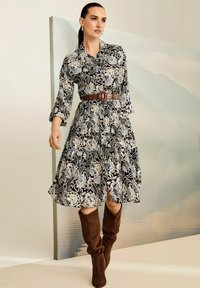 Frau in einem schwarzen Blumenkleid mit einem braunen Gürtel und knielangen braunen Wildlederstiefeln, die vor einem gedämpften Hintergrund mit Bergen steht.