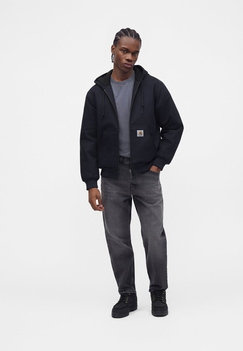 Joven con cabello trenzado que lleva una sudadera negra con cremallera, camisa gris, vaqueros negros desgastados y zapatos negros, de pie frente a un fondo simple.
