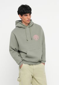 Olijfgroene hoodie van zacht katoen, met een voorzak in kangoeroestijl en een roze cirkelvormig logo op de linkerkant van de borst.