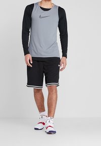 Camiseta deportiva gris sin mangas con mangas largas negras, combinada con pantalones cortos negros con rayas blancas y zapatillas deportivas blancas con detalles en rojo.