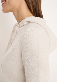 Haut de hoodie texturé beige clair, en tissu doux et duveteux. Il dispose d'un col côtelé et d'un cordon de serrage pour l'ajustement.