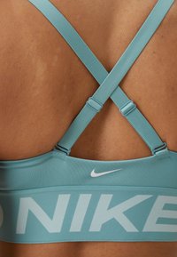 Plava sportska grudnjak s podesivim križnim trakama, s širim pojasom s "NIKE" logotipom u svijetlom fontu. Glatka tekstura, atletski dizajn.
