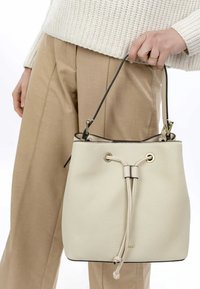 Bucket bag in textured cream leather with a black trim, adjustable drawstring closure, and gold-tone hardware. Handheld with a neutral outfit.