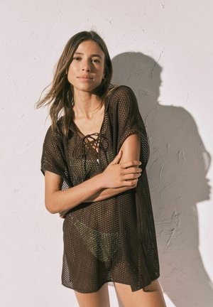 Vrouw met lang bruin haar die een losse, donkerbruine, opengebreide jurk draagt, staand tegen een gestructureerde witte muur met schaduw achter haar.