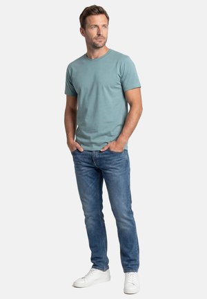 Mand står med hænderne i lommerne iført en blågrøn kortærmet t-shirt, blå jeans og hvide sneakers mod en ensfarvet baggrund.