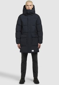 Sötétkék puffertürkiz magas gallérral és állítható kapucnival, két elülső zsebével és logóval a hemjén.