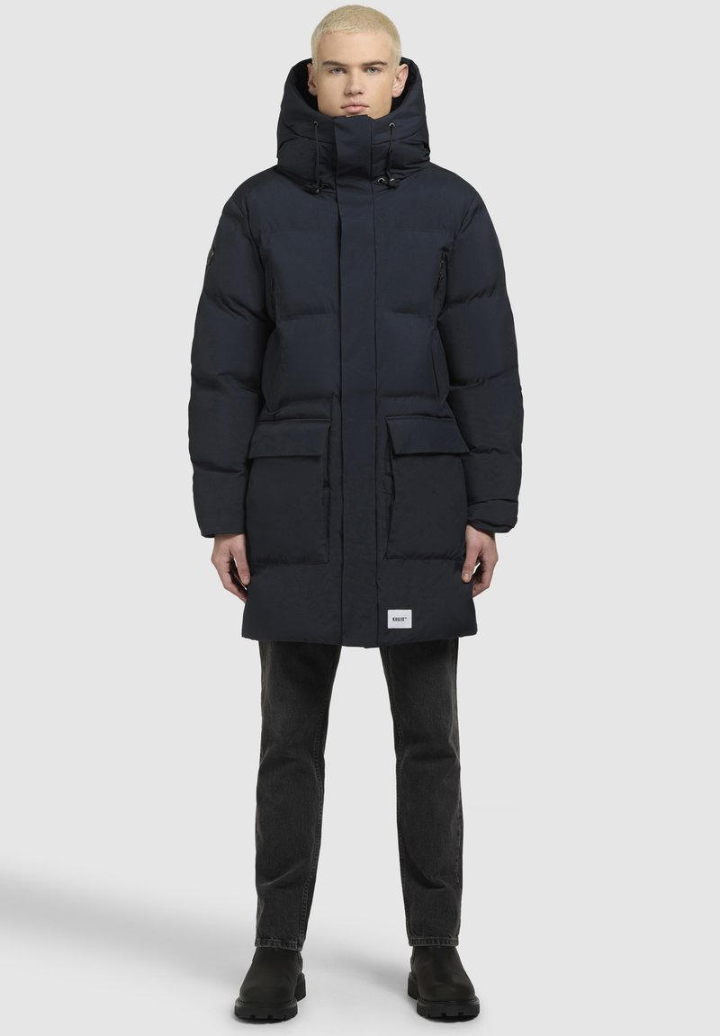 Sötétkék puffertürkiz magas gallérral és állítható kapucnival, két elülső zsebével és logóval a hemjén.