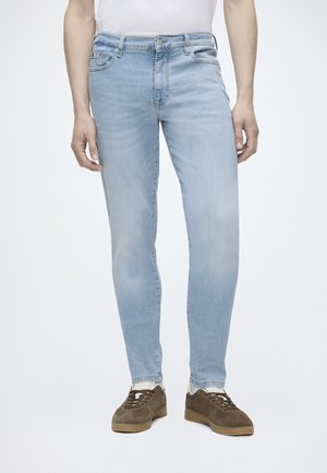 Person iført lyseblå slim-fit jeans, brune sneakers med hvide sokker og en hvid skjorte mod en ensfarvet hvid baggrund.