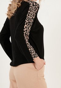 Černý top s dlouhým rukávem a leopardími potisky na rukávech. Měkká tkanina s volným střihem, spárovaná se světle barevnými kalhotami.