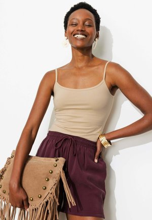 Femme souriante, les yeux fermés, portant un débardeur beige, un short bordeaux, des boucles d’oreilles et bracelets en or, tenant un sac à franges beige sur fond blanc.