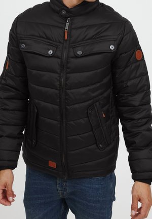 Schwarze Puffjacke mit einem Reißverschluss vorne, gerippten Bündchen und zwei Brusttaschen. Verziert mit braunen Lederakzenten und einem hohen Kragen.