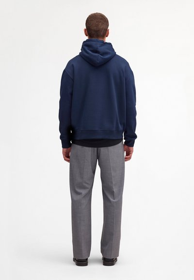 Mann steht mit dem Rücken zur Kamera, trägt einen dunkelblauen Kapuzenpullover, graue Anzughose und schwarze Schuhe vor einem schlichten weißen Hintergrund.