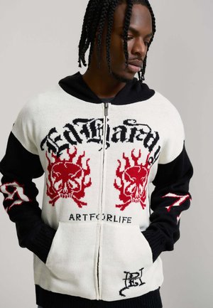 Jonge man met gevlochten haar, gekleed in een zwart-witte sweater met rits, voorzien van rode schedel- en vlamgrafieken en de tekst "ARTFORLIFE".