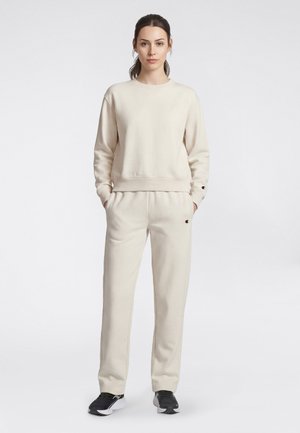 Frau steht und trägt einen beigefarbenen Pullover und passende Jogginghosen mit schwarz-weißen Turnschuhen vor hellem, einfarbigem Hintergrund.