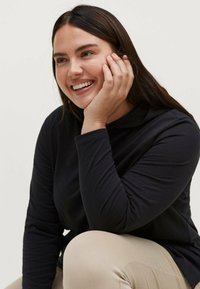 Sort langærmet top med en afslappet pasform og rund halsudskæring, parret med lyse beige bukser. Hånd i hudfarve hviler på kinden.