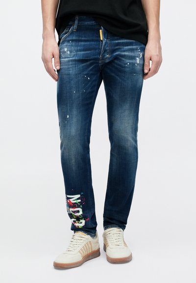 Τζιν μπλε denim με στενή γραμμή, διακοσμημένα με πιτσιλιές χρώματος και γραφικά κείμενα στο κάτω μέρος του αριστερού ποδιού. Συνδυασμένα με κρεμ αθλητικά παπούτσια.