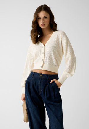 Femme aux cheveux bruns ondulés portant un cardigan crème texturé à col en V avec des boutons en forme de fleurs et un pantalon taille haute bleu foncé, tenant un sac beige.